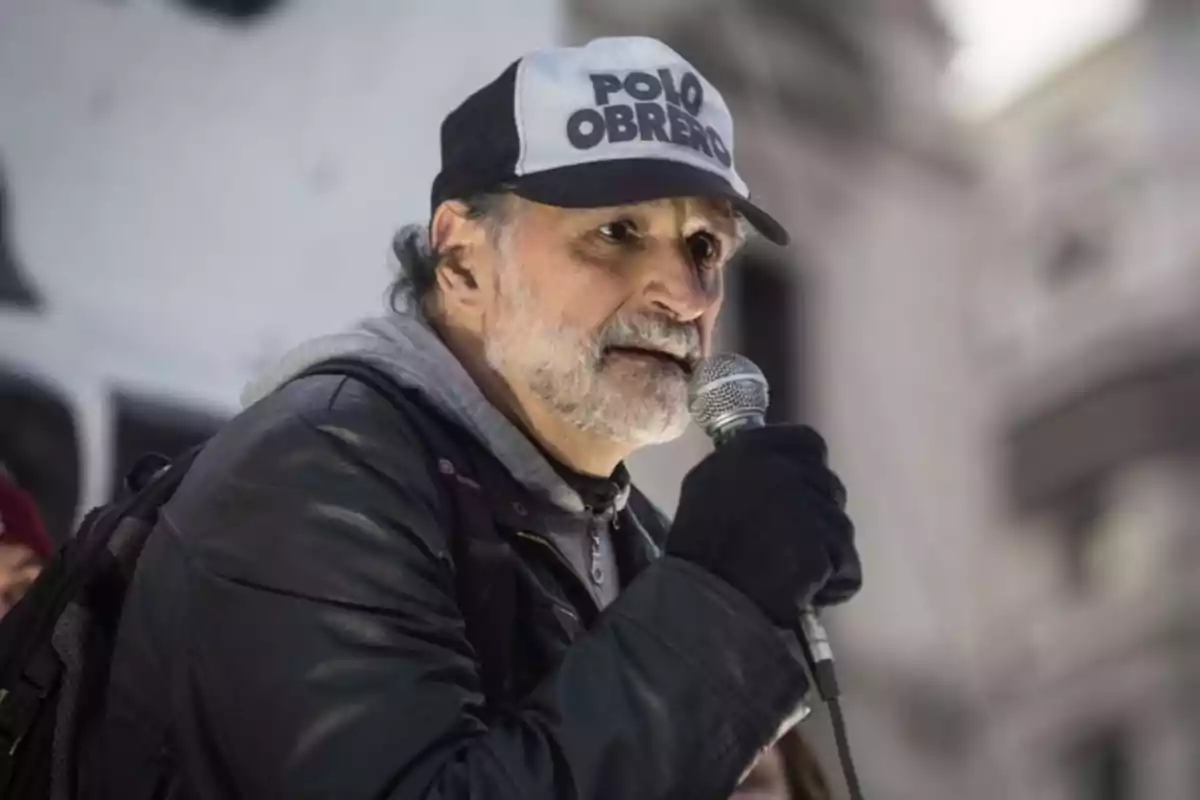 Un hombre con gorra y guantes sostiene un micrófono mientras habla en un evento al aire libre.