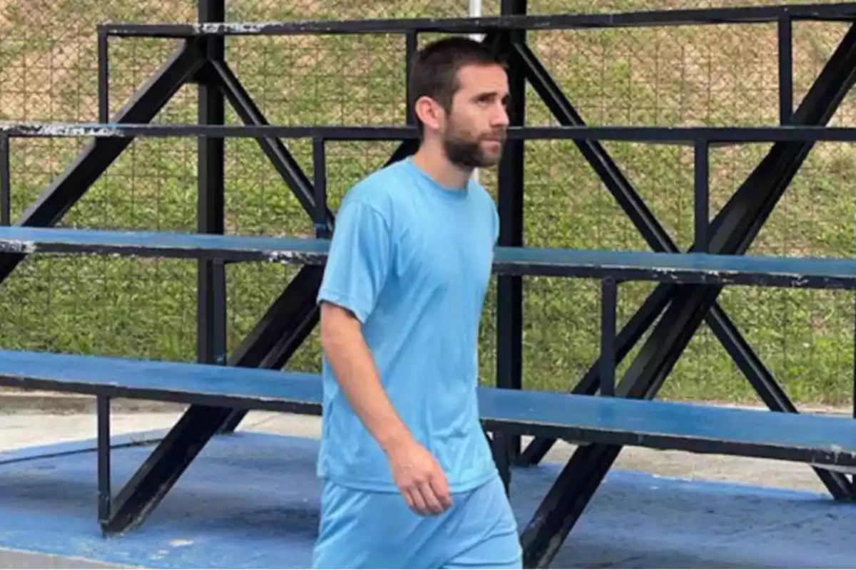 Hombre con camiseta azul caminando frente a unas gradas metálicas.