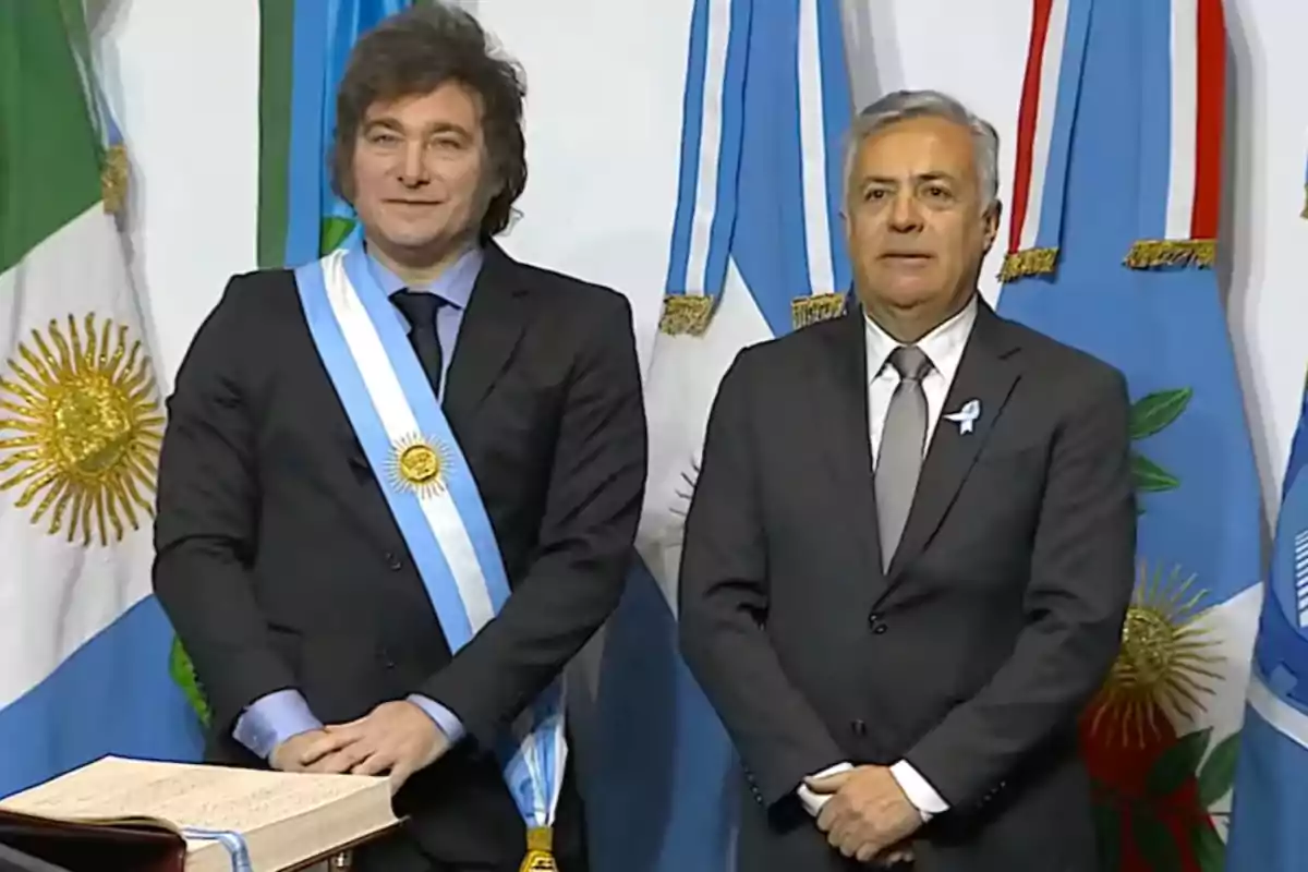 Dos hombres de pie con trajes formales y bandas presidenciales frente a banderas argentinas.