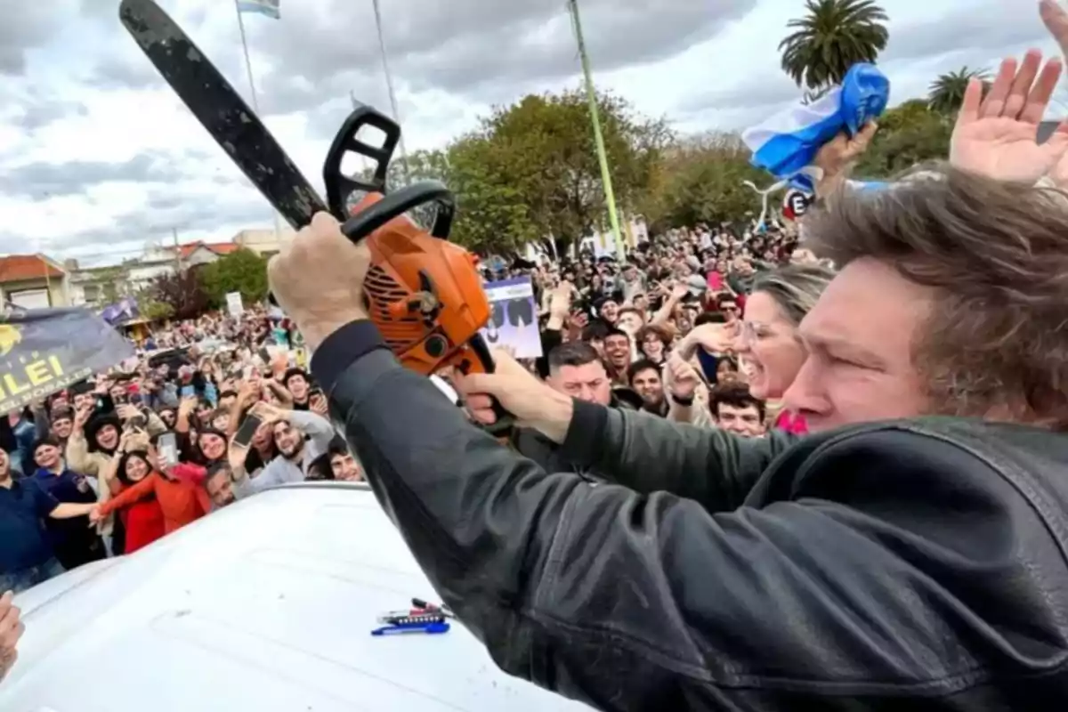 Una persona sostiene una motosierra en el aire mientras una multitud sonriente lo rodea en un evento al aire libre.