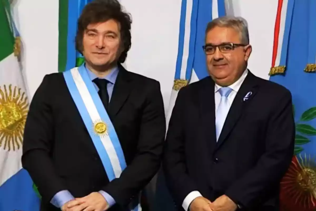 Dos hombres de traje posan frente a banderas argentinas, uno de ellos lleva una banda presidencial.