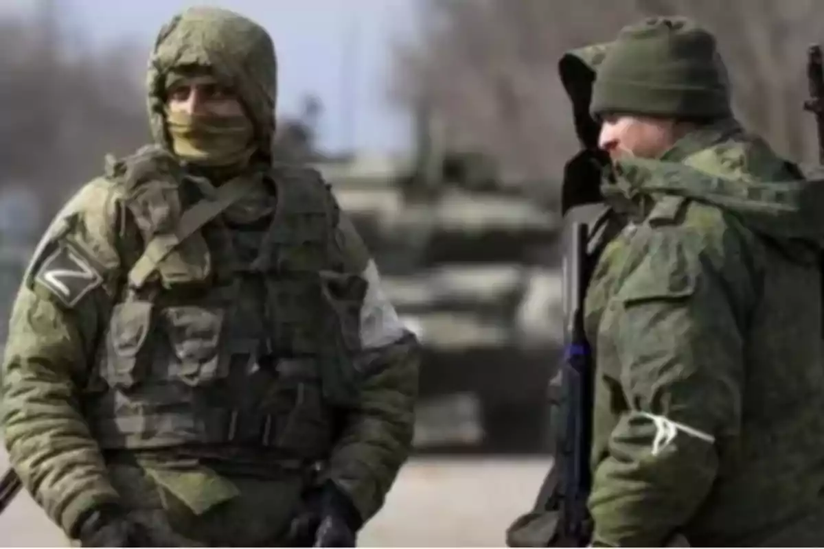 Dos soldados con uniforme de camuflaje verde y equipo militar, uno de ellos lleva una capucha y el otro un gorro, en un entorno al aire libre con vehículos militares de fondo.