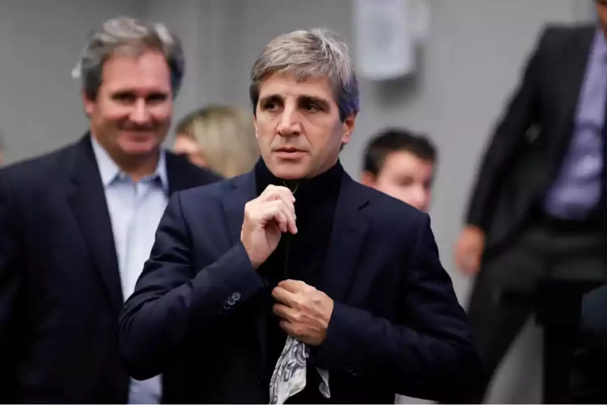 Un hombre de cabello canoso ajustándose el cuello de su chaqueta en un entorno formal con personas al fondo.