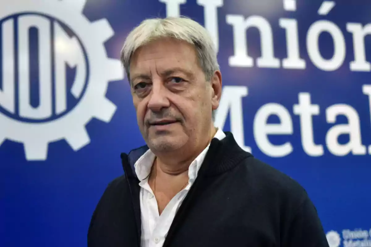 Hombre de cabello canoso con suéter negro frente a un cartel de la Unión Metalúrgica.