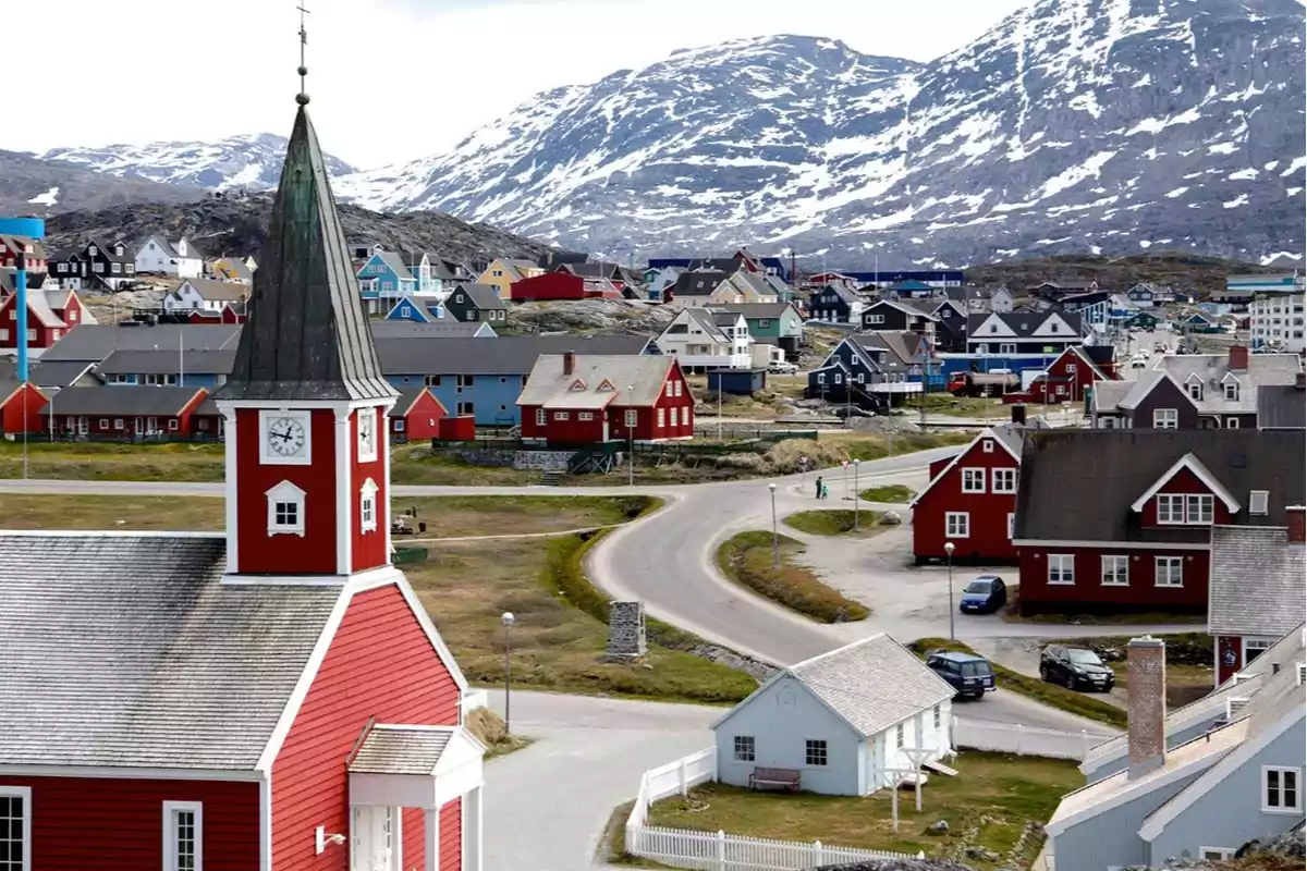 Una iglesia roja con un campanario y un reloj se encuentra en un pueblo con casas de colores y montañas nevadas al fondo.