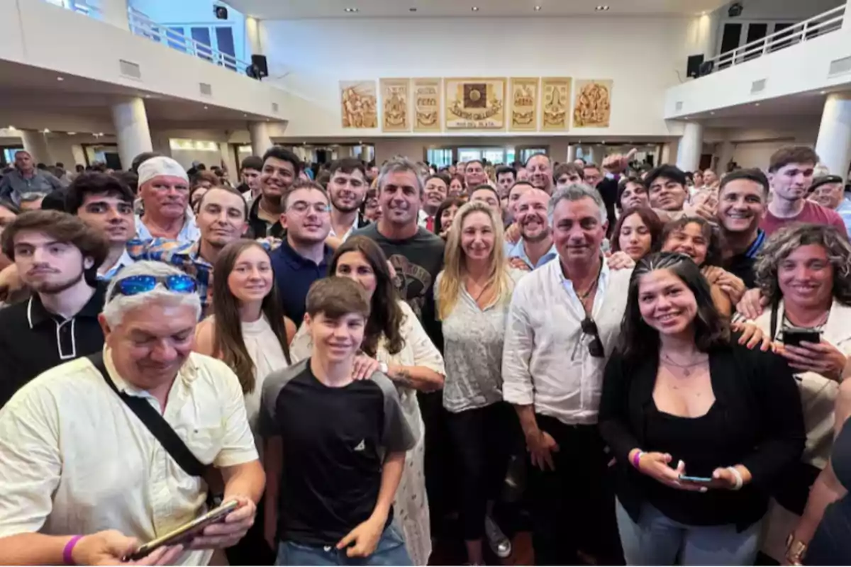 Un grupo numeroso de personas sonríe para una foto en un salón amplio con decoración en las paredes.