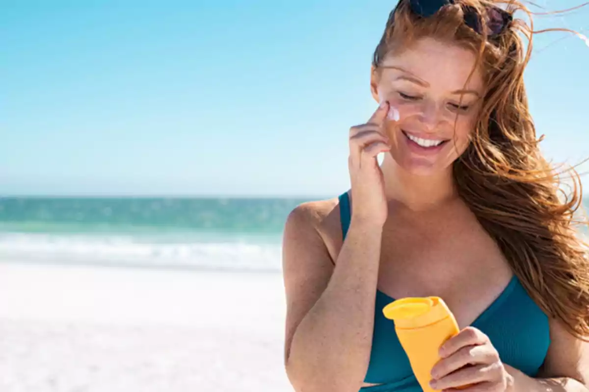 Mujer en la playa aplicándose protector solar en el rostro mientras sonríe.