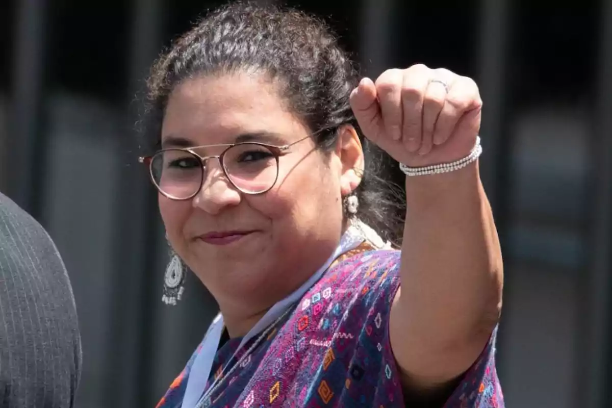 Una persona con gafas y aretes sonríe mientras levanta el puño, vistiendo una prenda colorida.