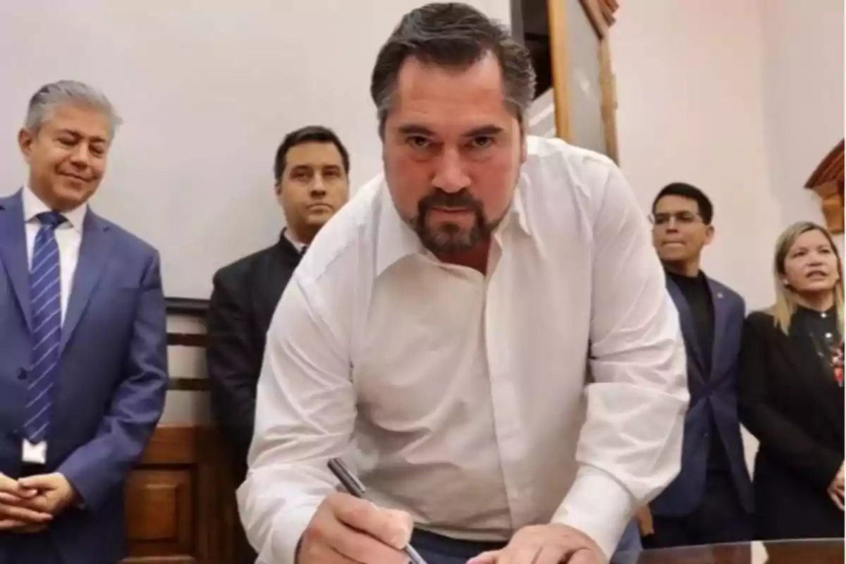 Un hombre firmando un documento rodeado de otras personas en una sala.