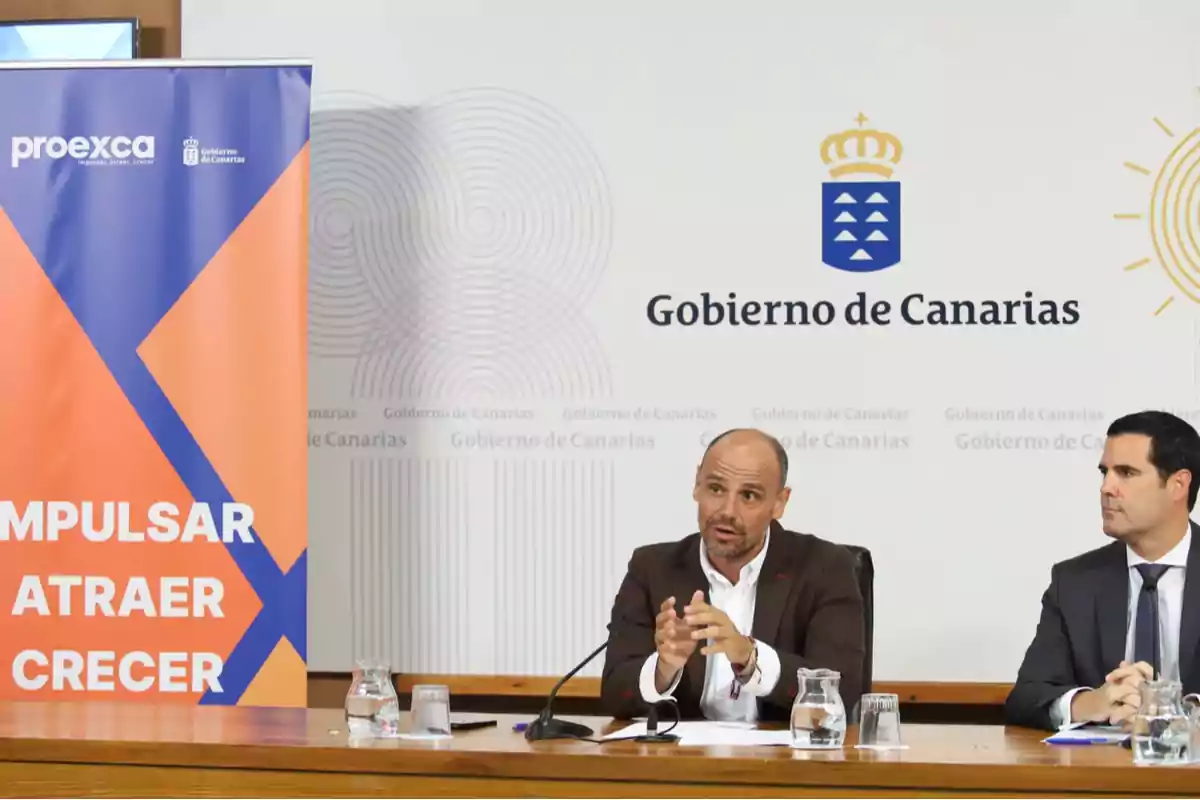 Dos hombres en una conferencia de prensa del Gobierno de Canarias con un cartel de Proexca que dice "Impulsar, atraer, crecer".