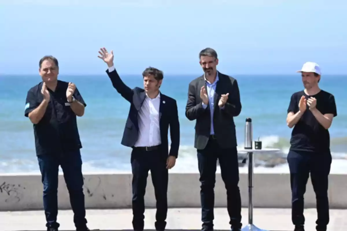 Cuatro personas de pie junto al mar, una de ellas levantando la mano mientras las otras aplauden.