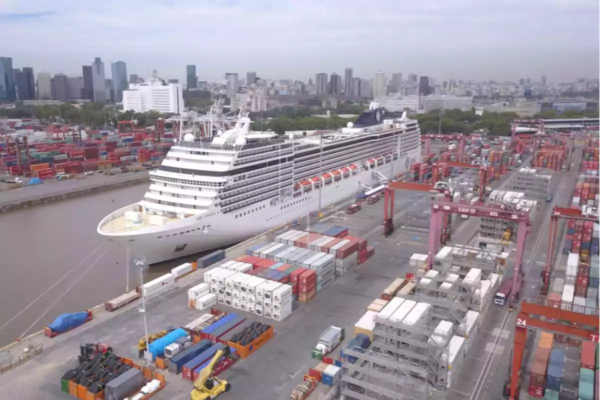 Un crucero atracado en un puerto rodeado de contenedores y grúas con una ciudad al fondo.