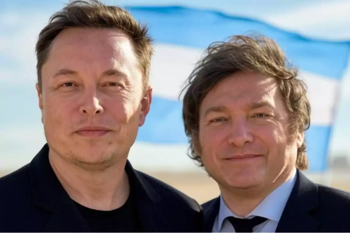 Dos hombres posando juntos frente a una bandera azul y blanca ondeando en el fondo.