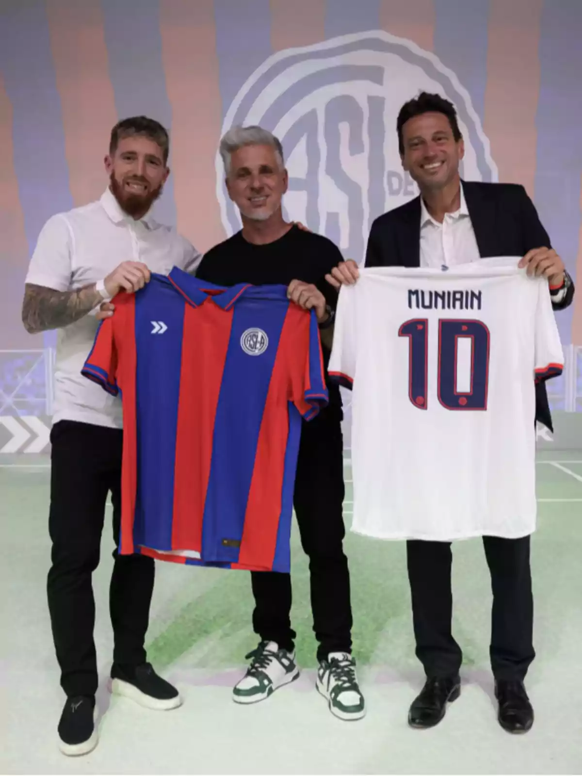 Tres personas posan sonriendo mientras sostienen camisetas de fútbol, una de rayas rojas y azules y otra blanca con el número 10 y el nombre Muniain.