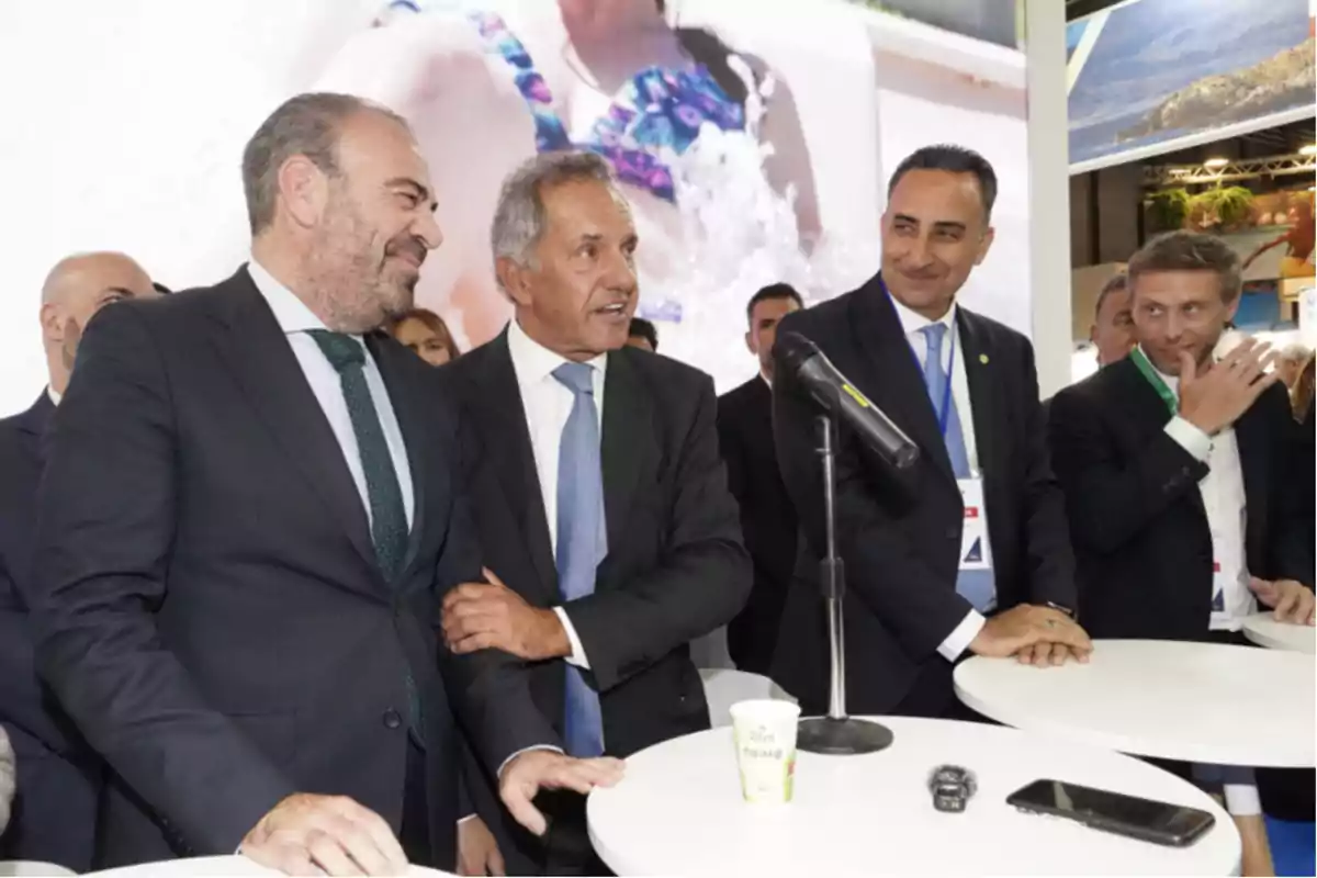 Un grupo de hombres en traje está reunido alrededor de una mesa blanca con un micrófono y un vaso de café, en un entorno de conferencia o evento.