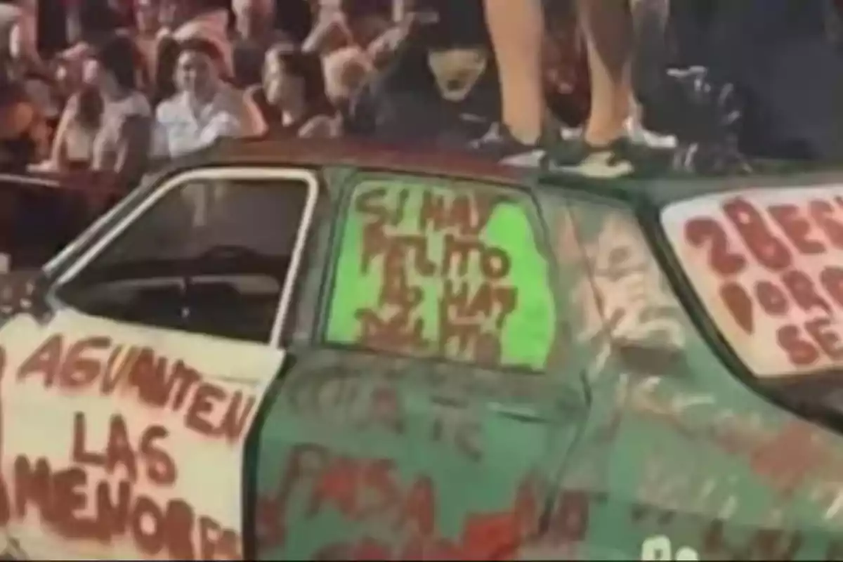 Un coche cubierto de grafitis con mensajes en las puertas y ventanas, rodeado de una multitud de personas.