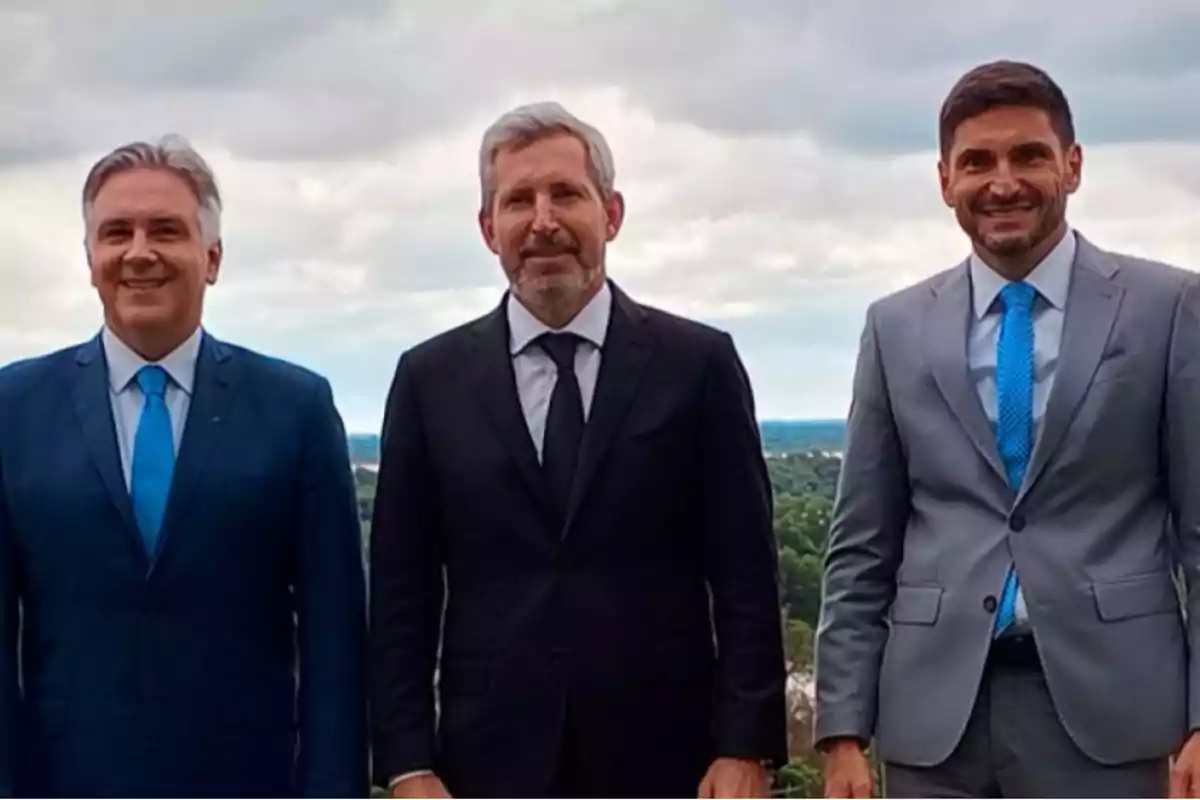 Tres hombres de pie con trajes formales y corbatas, posando al aire libre con un paisaje de árboles y cielo nublado de fondo.