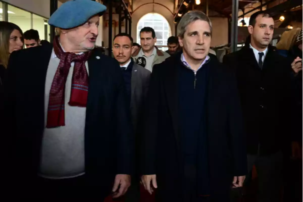 Un grupo de personas camina por un pasillo interior, destacando dos hombres al frente, uno con boina azul y bufanda roja, y el otro con cabello canoso y abrigo oscuro.