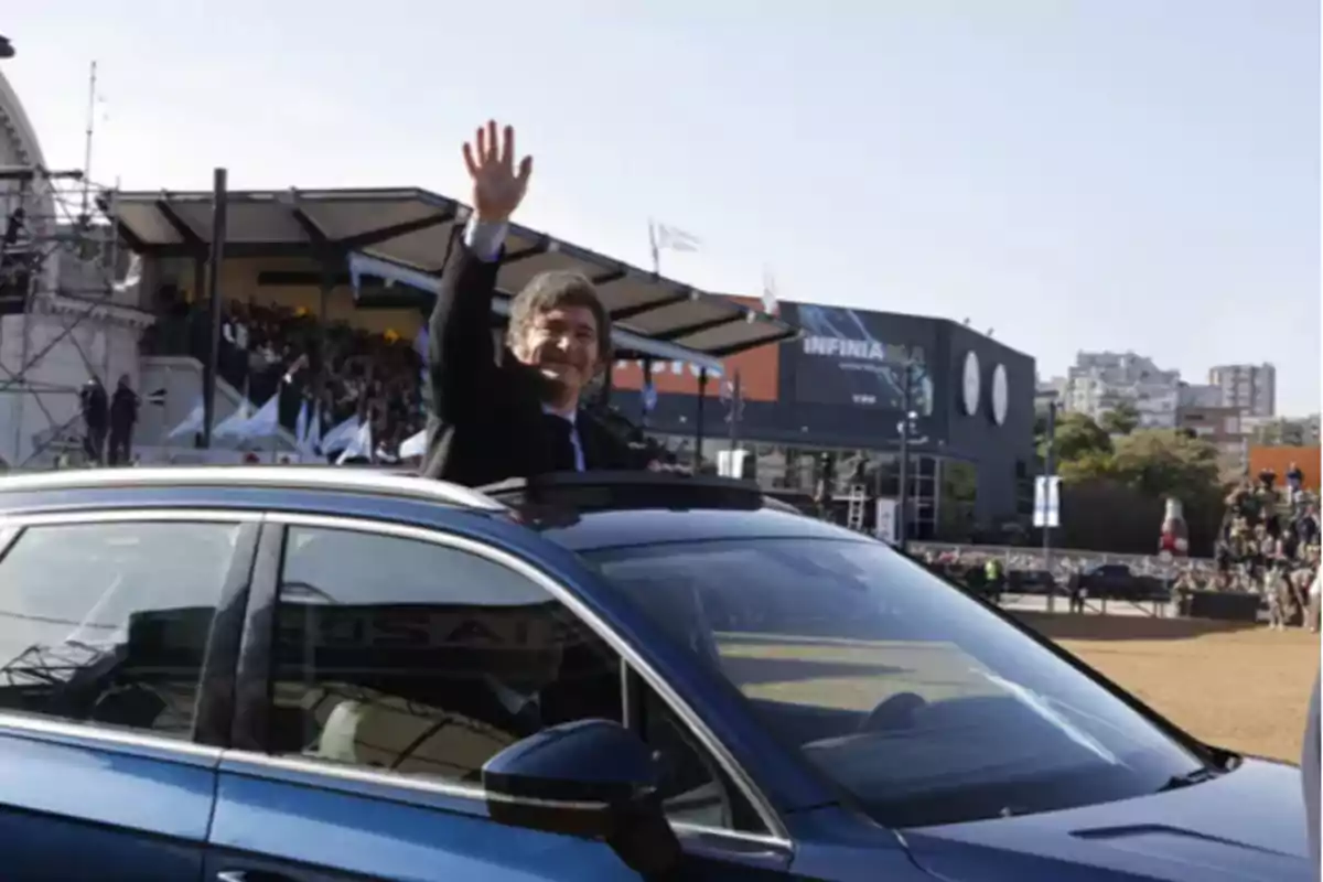 Una persona saludando desde el techo de un automóvil en un evento al aire libre con una multitud y banderas en el fondo.