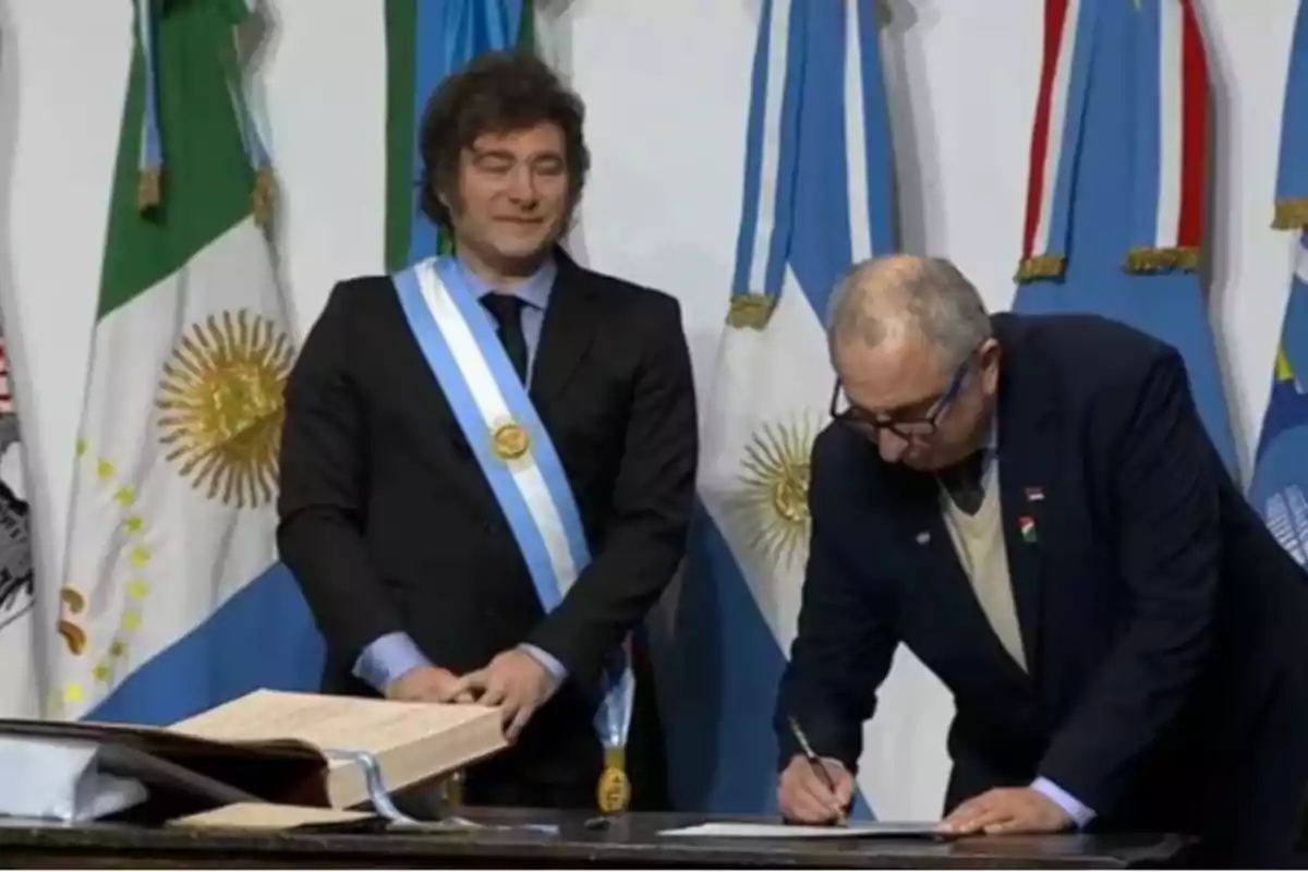 Dos personas en un evento formal, una de ellas lleva una banda presidencial y la otra está firmando un documento sobre una mesa, con banderas de fondo.