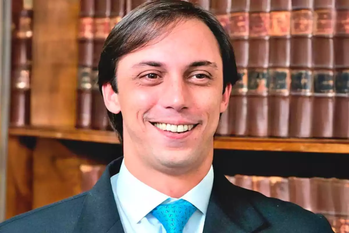 Un hombre sonriendo con una corbata azul frente a una estantería con libros.