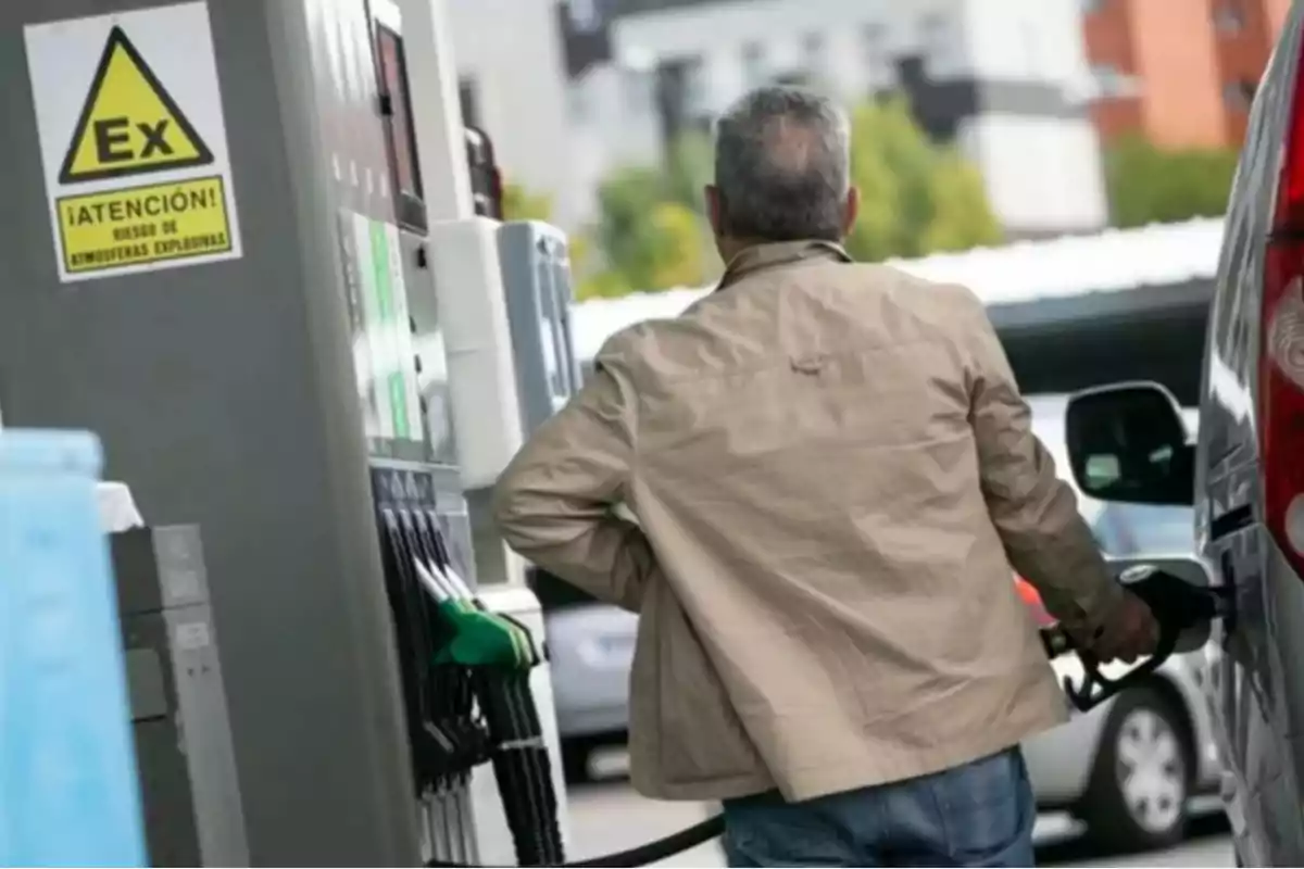 Un hombre de espaldas está llenando el tanque de su vehículo en una gasolinera mientras se observa un cartel de advertencia sobre riesgo de atmósferas explosivas.