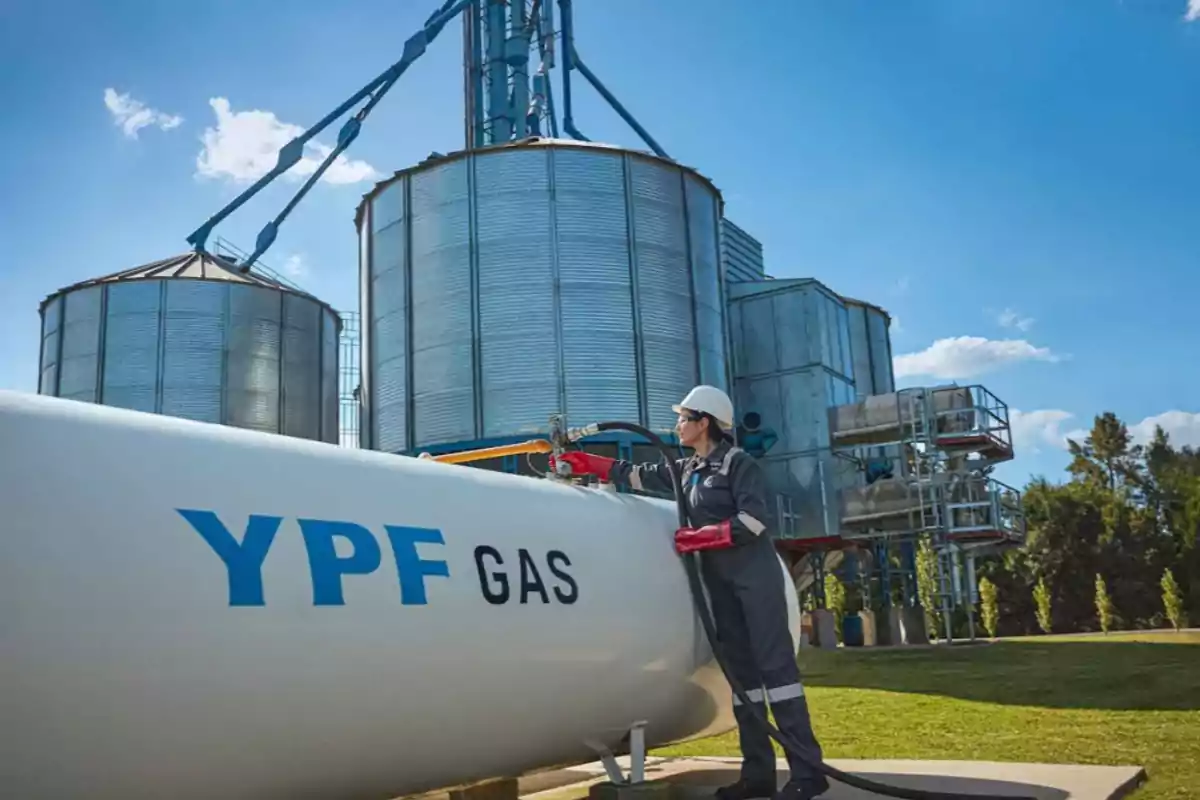 Una persona con casco y guantes rojos trabaja en un tanque de gas de YPF frente a silos metálicos bajo un cielo azul.