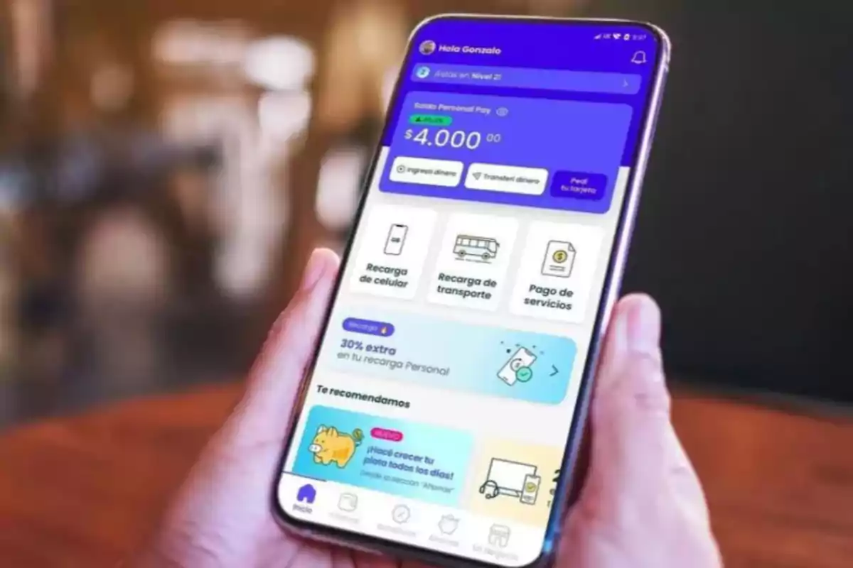 Una persona sostiene un teléfono móvil que muestra una aplicación de finanzas personales con opciones para recargar celular, transporte y pagar servicios.