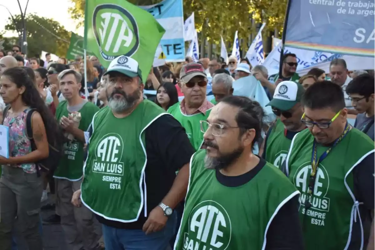 Un grupo de personas participa en una manifestación, algunas llevan chalecos verdes con el logo de ATE San Luis y hay banderas y pancartas en el fondo.