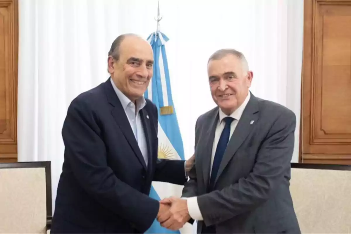 Dos hombres de traje se dan la mano frente a una bandera argentina en una sala con cortinas blancas.