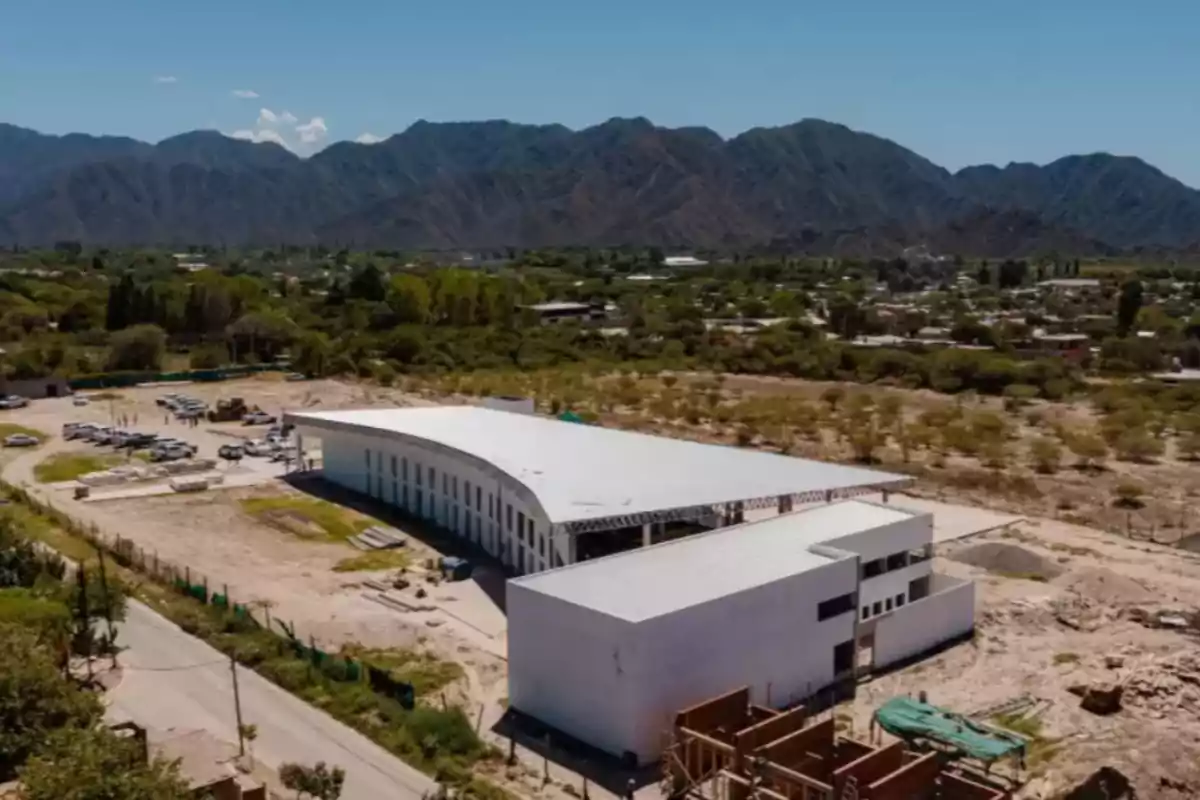 Vista aérea de un edificio moderno en construcción rodeado de vegetación y montañas al fondo.