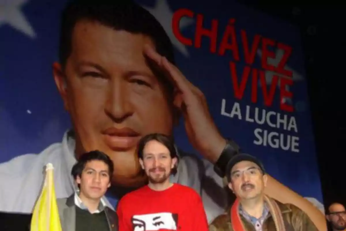 Tres personas posan frente a un cartel grande con la imagen de un hombre saludando y un texto que dice 