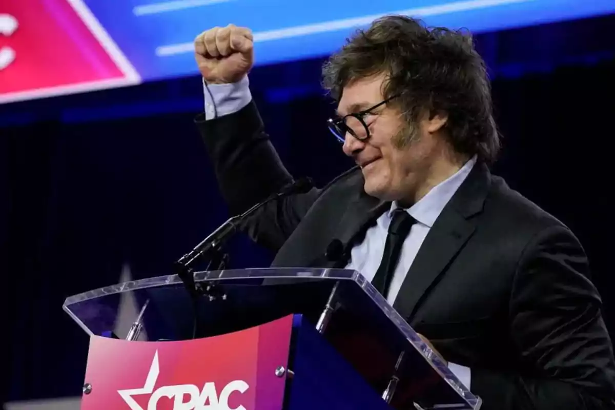 Un hombre con traje y gafas levanta el puño mientras habla en un podio con el logo de CPAC.