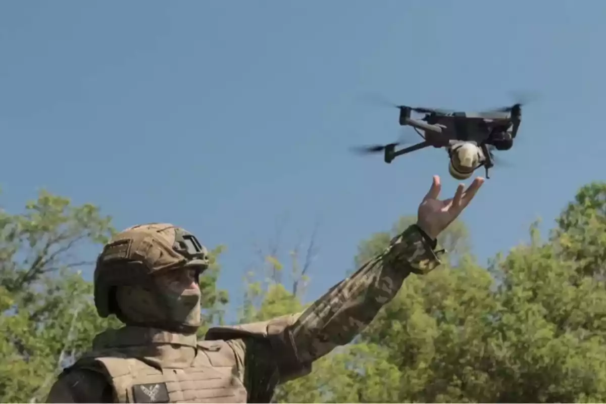 Un soldado en uniforme militar sostiene un dron en el aire con un fondo de cielo azul y árboles.