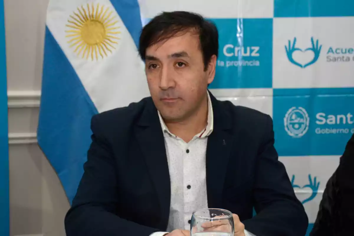 Un hombre con traje oscuro y camisa blanca está sentado frente a una bandera de Argentina y un fondo con logotipos.