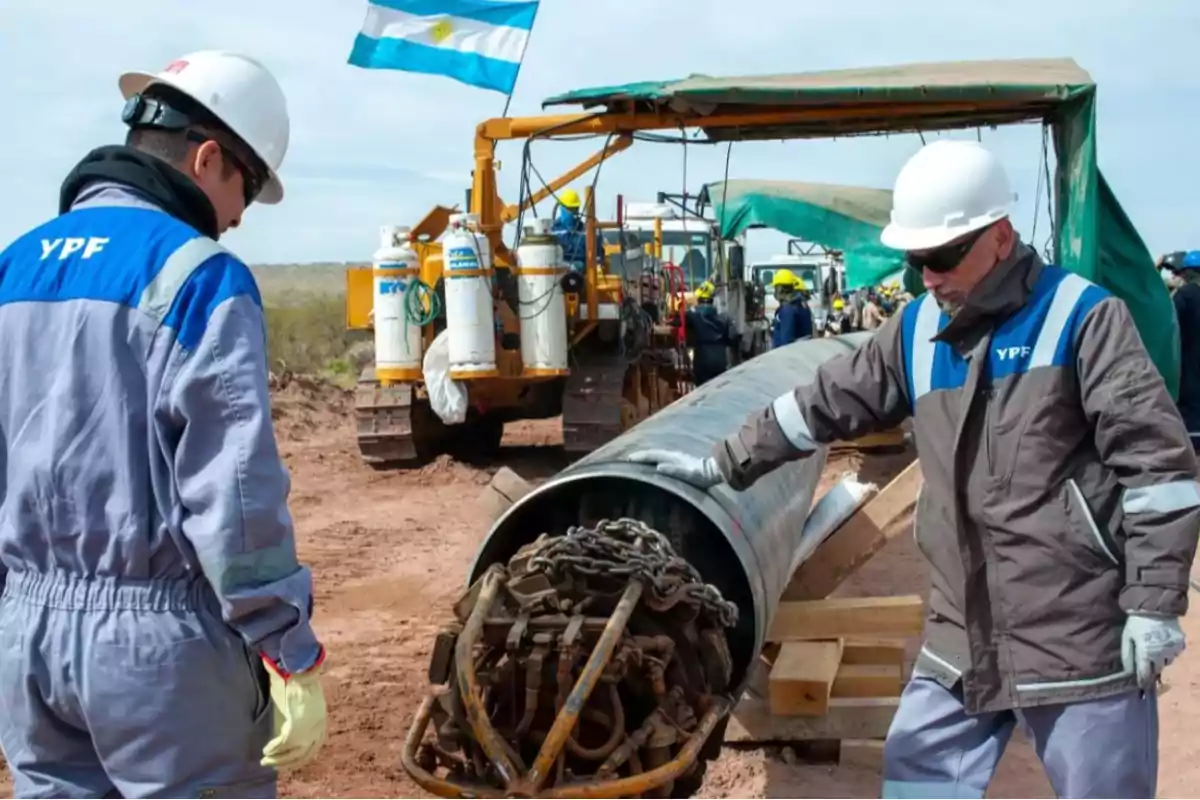 Trabajadores con uniformes de YPF inspeccionan un tubo grande en un sitio de construcción, con maquinaria y una bandera argentina al fondo.