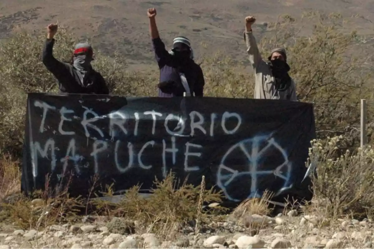 Tres personas con el rostro cubierto levantan el puño detrás de una pancarta negra que dice "Territorio Mapuche" en un paisaje árido.