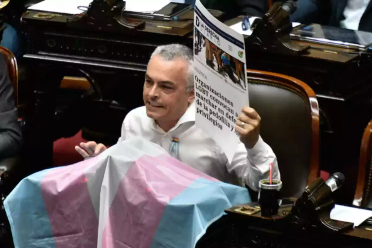 Un hombre sentado en un recinto parlamentario sostiene un cartel y una bandera de colores azul, rosa y blanco.