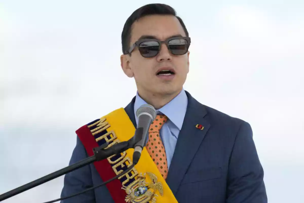 Un hombre con gafas de sol y traje azul habla frente a un micrófono, lleva una banda con colores rojo, amarillo y azul.