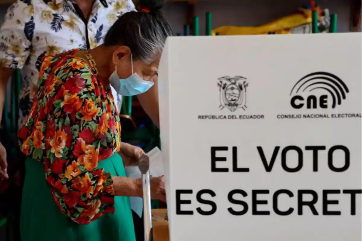 Una mujer con mascarilla y ropa colorida vota en una cabina electoral en Ecuador con un cartel que dice 