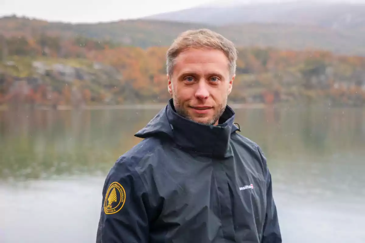 Un hombre con chaqueta oscura está de pie frente a un lago con un paisaje otoñal de fondo.