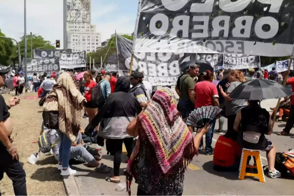 Un grupo de personas se reúne en una manifestación al aire libre con pancartas y banderas, algunas personas están sentadas mientras otras están de pie, y una mujer sostiene un abanico.