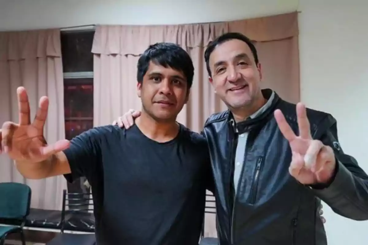 Dos personas posan juntas sonriendo y haciendo el signo de la paz con los dedos en una habitación con cortinas beige.