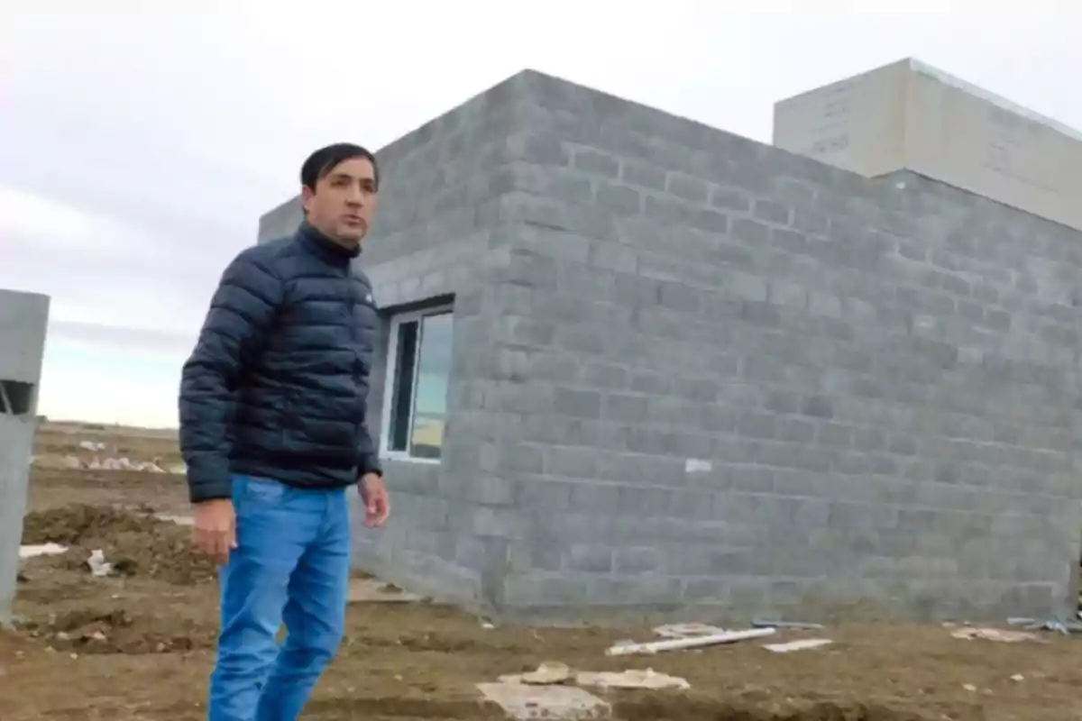 Un hombre con chaqueta negra y jeans de pie frente a una construcción de bloques grises en un terreno abierto.