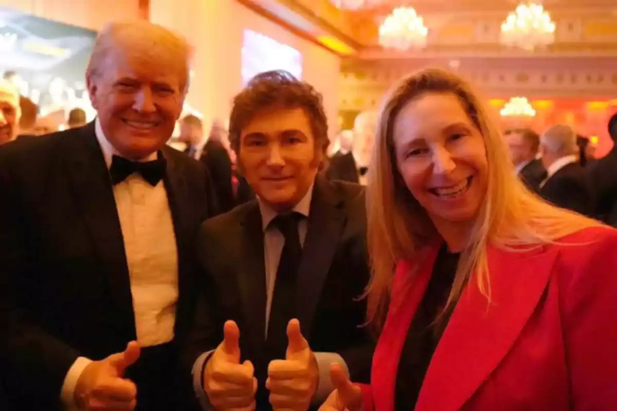 Tres personas posan sonriendo y levantando los pulgares en un evento formal con luces cálidas y candelabros al fondo.