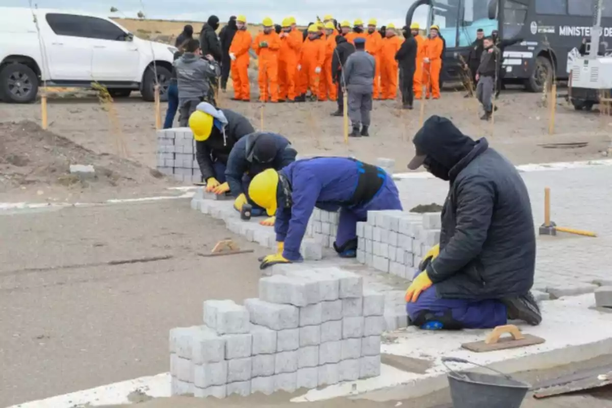 Trabajadores con cascos amarillos colocan adoquines en una obra mientras un grupo de personas con uniformes naranjas observa al fondo.