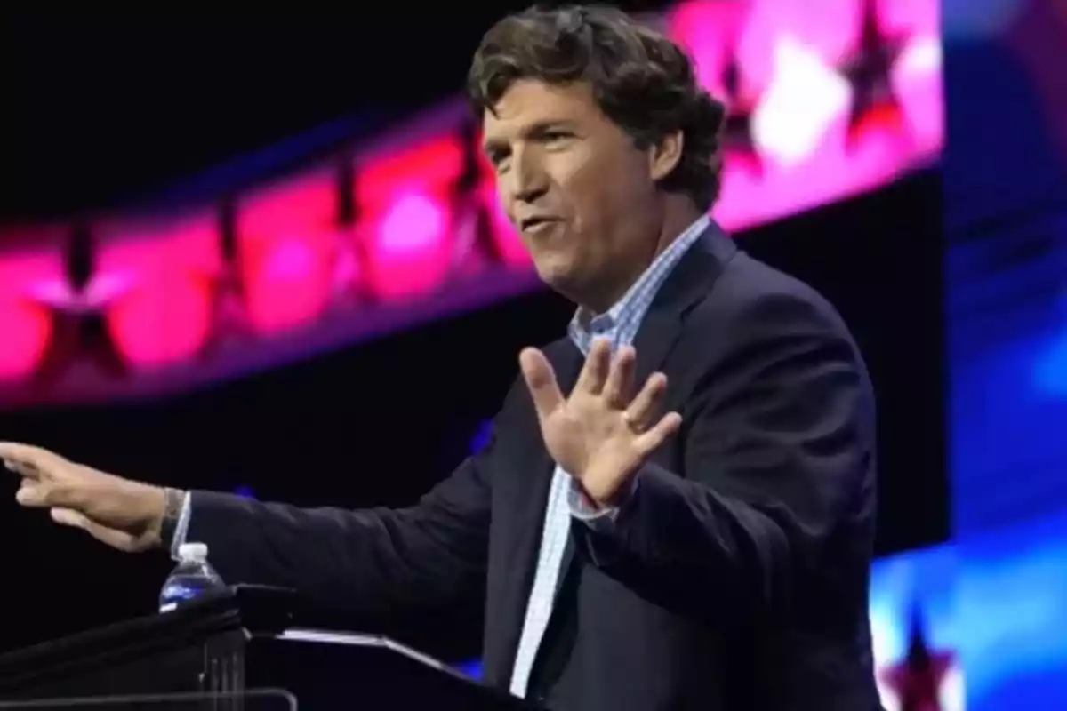 A man speaking on a stage with a background lit in pink and blue colors.