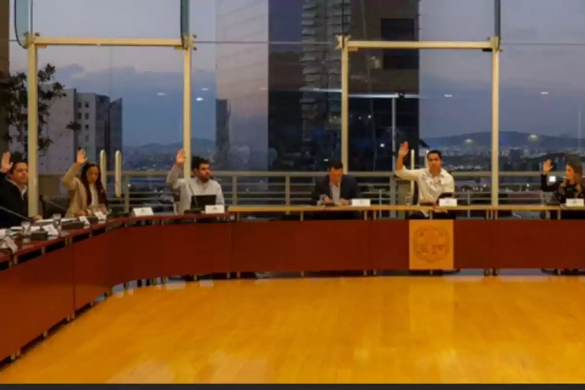 A group of people in a meeting room raise their hands while sitting around a table in a building with large windows showing a view of the city at sunset.