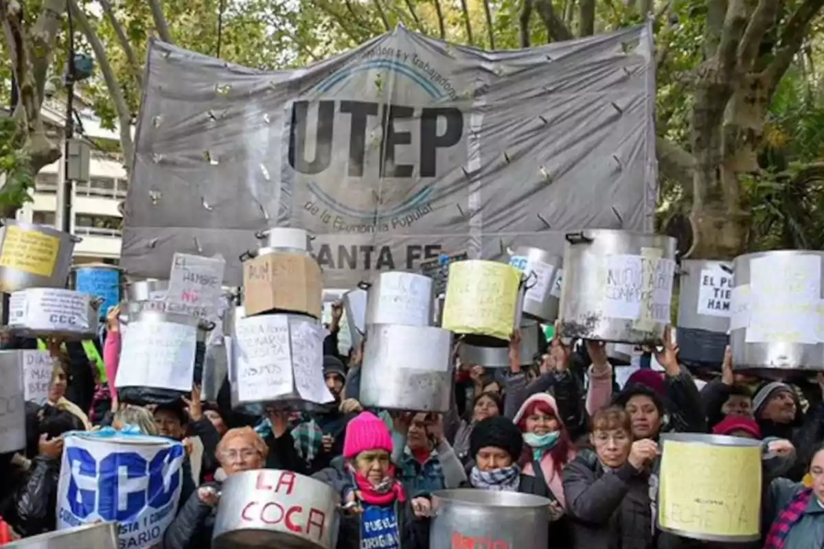 Un grupo de personas sostiene ollas y carteles en una manifestación al aire libre con un gran cartel de fondo que dice UTEP.