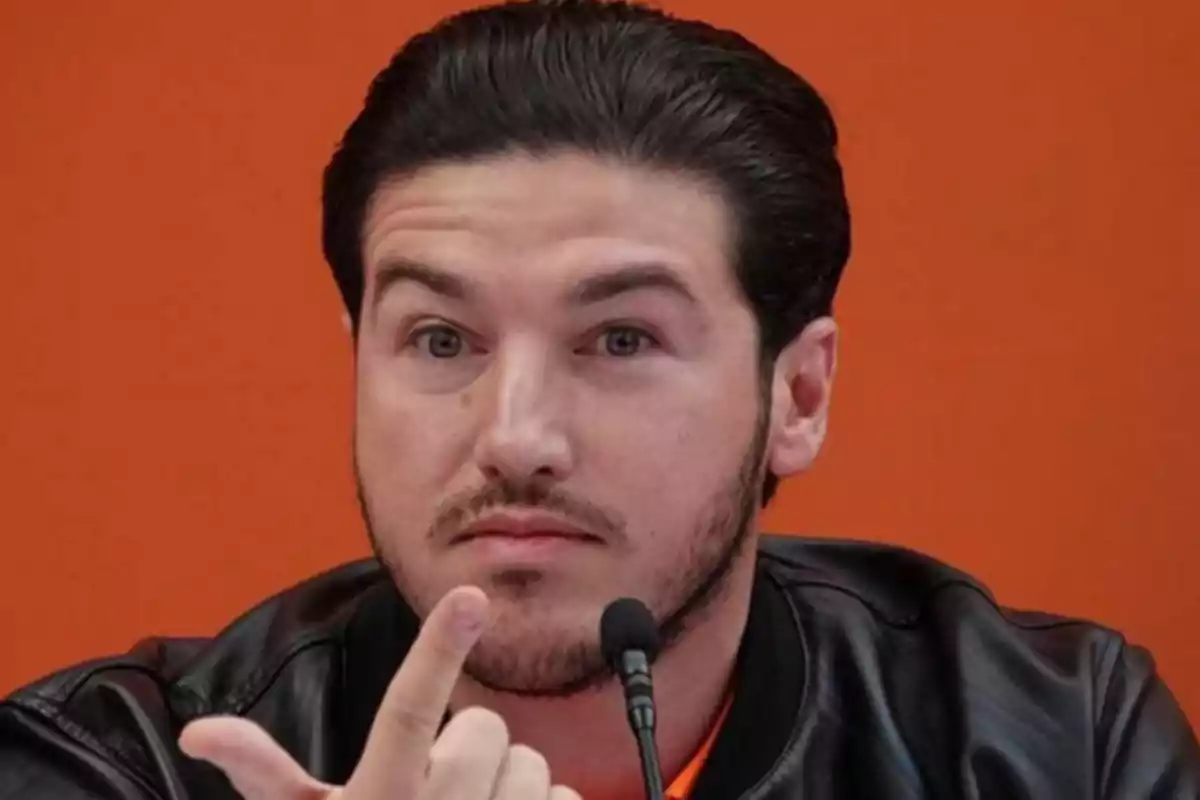 A man in a black leather jacket in front of an orange background speaking into a microphone.